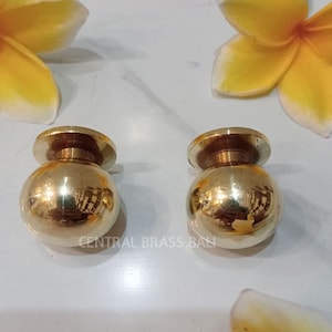 Könnte beinhalten: Zwei polierte Messing-Schrankknöpfe mit kugelförmigem Design. Die Knöpfe sind reflektierend, mit kreisförmiger Basis und abgerundeter Oberseite. Das Bild enthält auch gelbe und weiße Blumen sowie den Text "CENTRAL BRASS BALI".