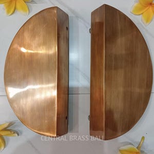 May include: Two bronze-colored, semi-circular door pulls. Each pull has a flat, rectangular section and a rounded, half-moon shape. The text "CENTRAL BRASS BALL" is printed at the bottom. Yellow flowers are placed around the door pulls.