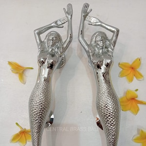 May include: A pair of silver mermaid door handles with raised scales and outstretched arms. The handles are shown against a white background.