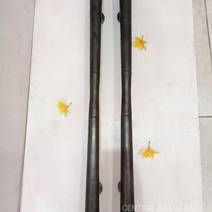 May include: A pair of dark bronze door pulls, each with etched detailing at the top and bottom. The cylindrical pulls have a subtle hourglass form. The text "CENTRAL BRASS BALI" is visible. Each pull is approximately 60 cm long.