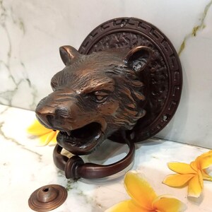Könnte beinhalten: Ein bronzefarbener Türklopfer aus Metall in Form eines knurrenden Tigerkopfes, montiert auf einer runden Rückplatte mit einem griechischen Mäander-Muster. Ein runder Ring hängt aus dem Maul des Tigers und ein kleines, separates Metallteil liegt daneben.