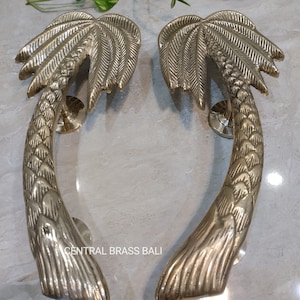 May include: Two brass palm tree door handles. The handles are detailed with textured fronds and trunks, and are mounted on circular bases. The handles are a warm gold color and are displayed on a light gray surface. The text "CENTRAL BRASS BALI" is visible.