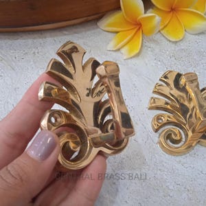 May include: Two polished brass wall hooks featuring a decorative leaf-shaped backplate with intricate scrollwork at the base and a single protruding hook arm. The metal surface is reflective and gold-toned, with visible screw holes for mounting.