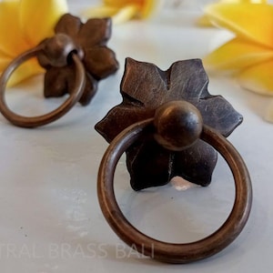 May include: Two bronze-colored flower-shaped cabinet pulls with circular handles. The pulls are set against a white surface, with yellow and white flowers in the background. The pulls have a vintage or antique appearance.