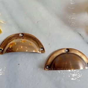 Puede incluir: Dos tiradores de latón antiguo con diseño semicircular. Cada tirador tiene tres orificios para tornillos para el montaje. Los tiradores son de un color dorado cálido, con un aspecto ligeramente envejecido. El fondo incluye flores amarillas.