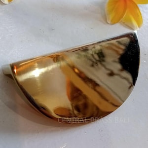 Könnte beinhalten: Ein polierter Messing-Schrankgriff in Halbmondform. Der Griff ist goldfarben und reflektiert das Umgebungslicht. Der Text "CENTRAL BRASS BALI" ist sichtbar. Gelbe und weiße Blumen befinden sich im Hintergrund.
