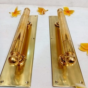 May include: Two polished brass door handles mounted on rectangular backplates. Each handle features a cylindrical grip with rounded ends and a smooth, reflective finish. The text "CENTRAL BRASS BALI" is printed in the bottom right corner.