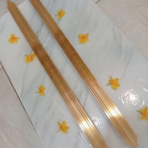 May include: Two gold-colored metal door handles with a decorative design. The handles are on a white marble surface with yellow flowers.