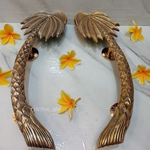 Op de afbeelding: Een paar goudkleurige deurgrepen in de vorm van palmbomen. De grepen zijn gebogen en hebben een gedetailleerd ontwerp.