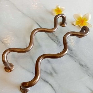 Puede incluir: Dos manijas de puerta onduladas de color bronce. Cada manija tiene una base circular y un extremo redondeado. Las manijas se exhiben sobre una superficie de mármol blanco, con flores amarillas y blancas en el fondo.