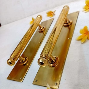 Pode incluir: Duas maçanetas de porta douradas com um design longo e cilíndrico e pontas arredondadas. Cada maçaneta é montada em uma placa retangular de latão com furos para parafusos. As placas estão posicionadas em uma superfície branca.