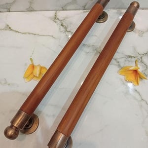 May include: Two wooden door handles with brass detailing. The handles are a warm brown colour with a cylindrical design and rounded ends. The brass hardware includes circular mounting plates and decorative end caps. The text "CENTRAL BRASS BALI" is visible.