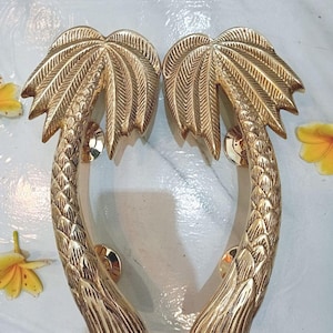 May include: A pair of gold-coloured palm tree door handles. The handles are made of metal and feature detailed palm fronds and trunks. The handles are displayed on a white surface with yellow flowers.
