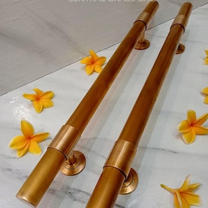 May include: Two cylindrical brass handrails with circular mounting plates and a band detail. The text "CENTRAL BRASS BALL" is at the top. The handrails are surrounded by yellow flowers. The overall design is a warm, golden tone.