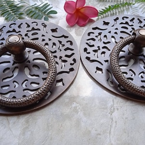 May include: Two round, ornate, brass door handles with intricate cut-out designs. Each handle features a ring pull with a textured surface.