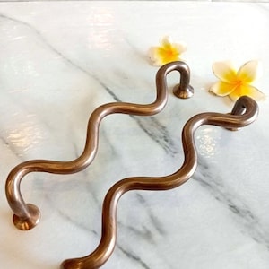 May include: A pair of bronze-coloured, serpentine cabinet handles. Each handle has a circular base and a curved, wavy design. They are displayed on a white marble surface, with yellow and white flowers in the background.