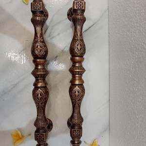 Op de afbeelding: Twee sierlijke bronzen deurklinken met bloemen- en geometrische patronen. De handgrepen hebben een verticale, langwerpige vorm met gedetailleerde gravures en een warme, antieke afwerking. De tekst "CENTRAL BRASS BALI" is zichtbaar onderaan.