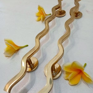 May include: Two gold-toned door handles with a wavy design. The handles are made of metal and have a round base. The handles are on a white surface with yellow flowers.