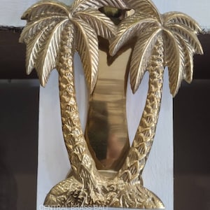 Może przedstawiać: Złoty mosiężny klamka do drzwi w kształcie dwóch palm. Klamka jest zamontowana na białych drzwiach. Tekst "CENTRAL BRASS BALI" jest widoczny u dołu klamki.