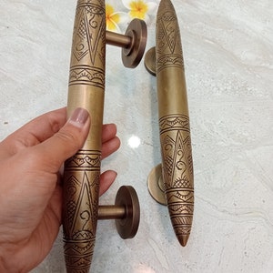 May include: Two ornate brass door handles with intricate geometric designs. The handles are long and slender, with a pointed tip. They have a polished finish and are mounted on round bases.