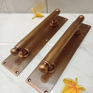 May include: Two antique bronze door pull handles. Each handle has a rectangular backplate with rounded ends and a cylindrical grip with a spherical end. The handles are made of bronze and have a weathered finish. The text "CENTRAL BRASS BALI" is visible.