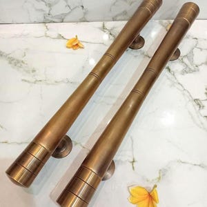 Op de afbeelding: Twee lange, cilindrische deurgrepen van geborsteld messing met een warme, metalen afwerking. Elke greep heeft een reeks horizontale groeven bij de uiteinden en is gemonteerd op ronde voetstukken. De tekst CENTRAL BRASS BALI is zichtbaar.