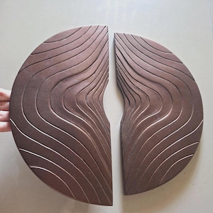 Puede incluir: Dos paneles decorativos de pared semicirculares con un acabado en bronce metálico y una superficie texturizada con surcos profundos, fluidos y ondulados. Las piezas están colocadas lado a lado, reflejándose para crear un patrón orgánico.