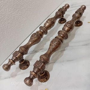 May include: Two bronze door handles with detailed floral and geometric patterns. The handles have a cylindrical design with decorative detailing and circular mounting plates. The text "CENTRAL BRASS BALI" is visible on the surface.