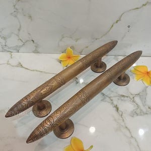 May include: A pair of bronze door handles with detailed engraved designs. The handles are long and cylindrical, with a pointed end. They are mounted on circular bases. The background is a white marble surface with yellow flowers.