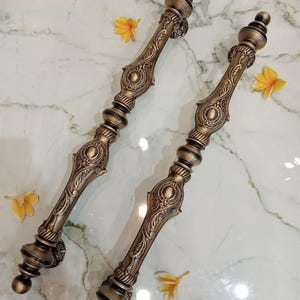 May include: Two decorative bronze door handles with detailed floral and geometric patterns. The handles are presented on a marble surface, adorned with yellow flowers. The text "CENTRAL BRASS BALI" is visible.