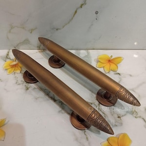 May include: Two bronze-colored door handles with a cylindrical design and decorative, pointed ends. Each handle is mounted on a circular base. The handles are displayed on a white marble surface, with yellow flowers scattered around.