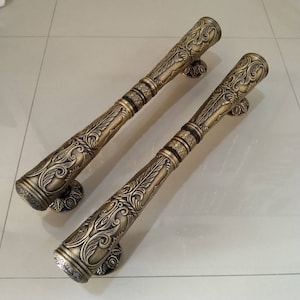 Puede incluir: Un par de tiradores de puerta ornamentados, de color dorado, con intrincados diseños florales. Los tiradores son largos y delgados, con un extremo redondeado y una base decorativa.