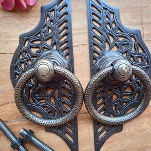 Puede incluir: Un par de tiradores de puerta de metal negro adornados con diseños intrincados y manijas de anillo. Los tiradores tienen un acabado desgastado y están montados en una superficie de madera.