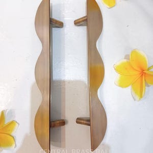 May include: Two brass door handles with a wavy design and wooden grips. The handles are a light brown color with a slight golden hue. The text "CENTRAL BRASS BALI" is visible at the bottom. Yellow flowers are scattered around the handles.