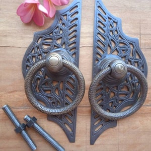 Pode incluir: Duas puxadores de porta de metal preto adornadas com alças de anel. As puxadores têm um design floral decorativo e são montadas numa superfície de madeira.