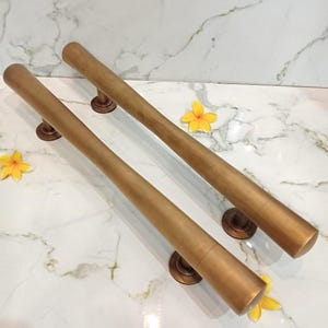 Könnte beinhalten: Zwei zylindrische Türgriffe in Bronzefarbe mit kreisförmigen Montageplatten. Die Griffe sind diagonal auf einer weißen Marmoroberfläche positioniert, mit gelben Blumen darum herum. Sie haben eine glatte, metallische Oberfläche.