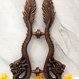 May include: A pair of antique bronze-colored brass door handles featuring detailed dragon motifs with scaled bodies and fin-like tops. The handles are connected by two central cylindrical mounts. The text CENTRAL BRASS BALI is visible at the top.