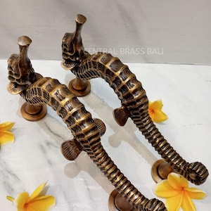 Könnte beinhalten: Zwei bronzefarbene Türgriffe in Form von Seepferdchen. Die Griffe sind mit detaillierten Schuppen und Flossen versehen und auf runden Sockeln montiert. Das Bild enthält auch gelbe Blumen und den Text "CENTRAL BRASS BALI".