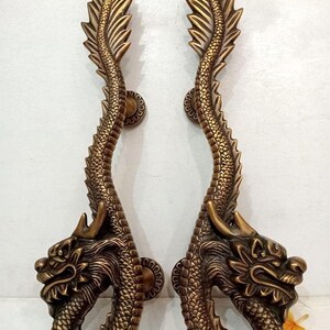 Könnte beinhalten: Zwei bronzefarbene Türgriffe in Drachenform. Die Griffe sind aufwendig gestaltet und weisen Drachenköpfe an der Basis und einen geschuppten Körper auf, der sich nach oben krümmt. Der Text "CENTRAL BRASS BALI" ist im Hintergrund sichtbar.