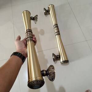 Op de afbeelding: Twee sierlijke gouden deurgrepen met decoratieve details. De grepen zijn lang en cilindrisch met een afgerond uiteinde. Ze hebben een gepolijste afwerking en ingewikkelde ontwerpen.