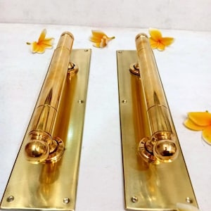 May include: Two polished brass door handles with rectangular backplates, each featuring a cylindrical grip and rounded ends. The hardware is metallic gold in color and includes visible screw holes for mounting. The text CENTRAL BRASS BALI is present.