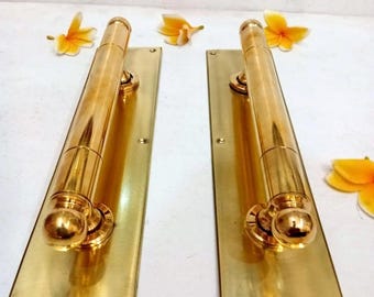 Könnte beinhalten: Zwei polierte Messing-Türgriffe mit rechteckigen Rückplatten, jeweils mit einem zylindrischen Griff und abgerundeten Enden. Die Beschläge sind metallisch goldfarben und haben sichtbare Schraublöcher. Der Text CENTRAL BRASS BALI ist zu sehen.