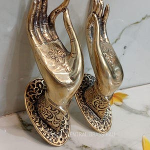 May include: Two brass-colored hand sculptures with intricate detailing. The hands are in a gesture of offering or blessing, mounted on decorative oval bases. The sculptures have a polished finish and are likely for home decor.