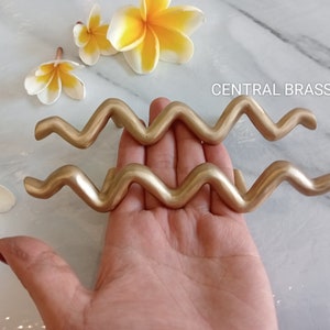 May include: Two gold-coloured metal drawer pulls shaped like the astrological symbol for Aquarius. The pulls are on a hand against a white marble background. The text "CENTRAL BRASS BALI" is visible in the image.
