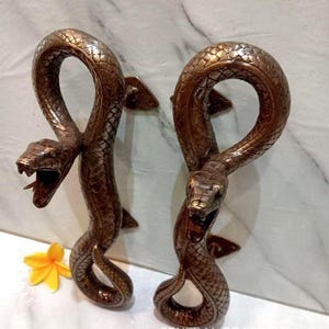 May include: A pair of bronze-colored metal door handles shaped like coiled snakes with textured scales and open mouths. The handles feature mounting brackets and the text CENTRAL BRASS BALI is visible at the bottom right of the image.