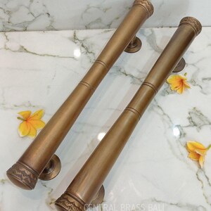 May include: Two bronze-colored door handles with decorative ends and circular mounting plates. The handles are cylindrical with etched detailing near the ends. The text "CENTRAL BRASS BALI" is visible.