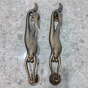 Puede incluir: Dos tiradores de puerta de sirena de oro antiguo con detalles intrincados. Las sirenas están representadas con cabello y colas flotantes, y sus rostros son serenos. Los tiradores están montados sobre un fondo blanco.