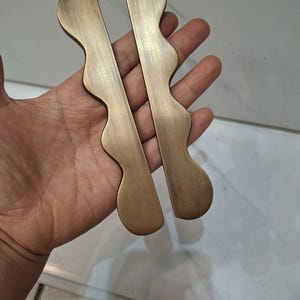 May include: Two bronze-colored cabinet pulls with a wavy design. The pulls have a rectangular shape with a curved edge and a smooth, metallic finish. They are likely used for drawers or cabinet doors.