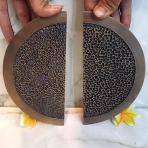 Puede incluir: Dos tiradores de puerta metálicos semicirculares con acabado en bronce oscuro y una superficie texturizada y picada. Cada tirador tiene un borde exterior liso y plano y una sección interior curva con un relieve profundo de panal irregular.