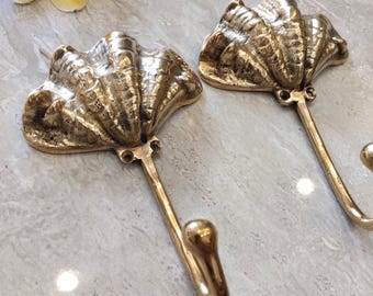Può includere: Due ganci da parete in ottone color oro a forma di conchiglie testurizzate. Ogni gancio presenta una parte superiore a conchiglia larga e scanalata con un braccio metallico curvo verso il basso, che termina con un pomello arrotondato. Il testo CENTRAL BRASS BALI è visibile.